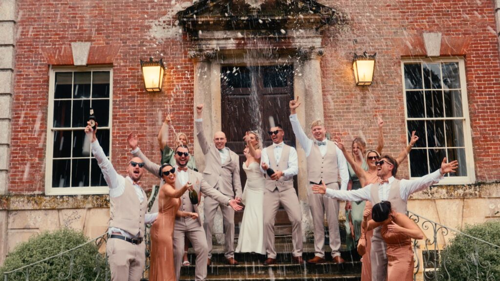 Bridal party celebrate with a champagne spray alongside the bride and groom on their wedding day. Captured by cool uk wedding videographer HC Visuals.