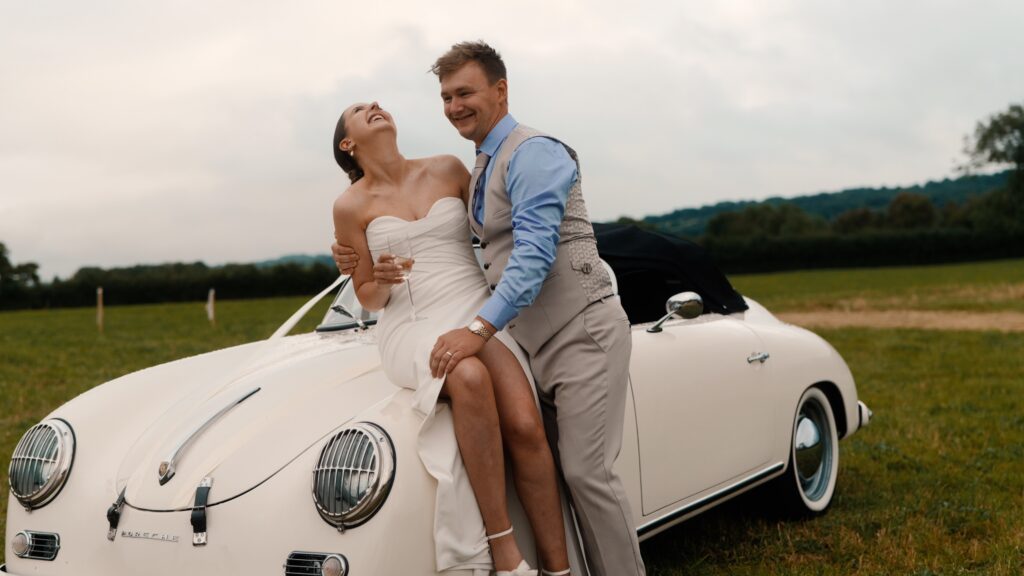 A couple sharing a fun moment with each other and their classic car during their wedding day in the Cotswolds. 